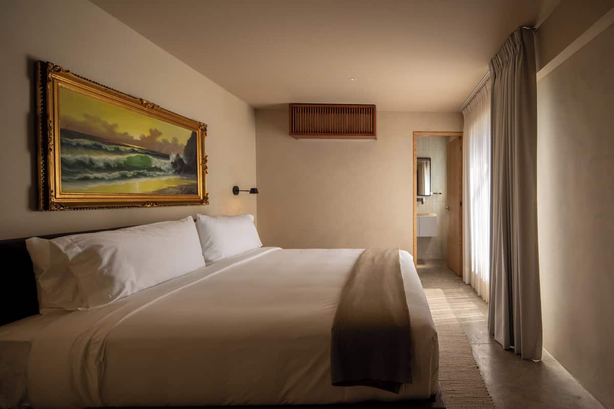 Une chambre d’hôtel avec un grand lit, un tableau au mur, une grille d’aération en bois et une vue sur la salle de bain à travers une porte.