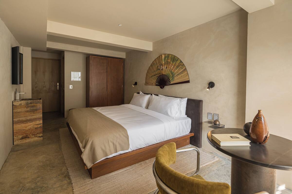 Une chambre d’hôtel moderne avec un grand lit, une armoire en bois, un bureau agrémenté d’un vase et un ventilateur fixé au mur.