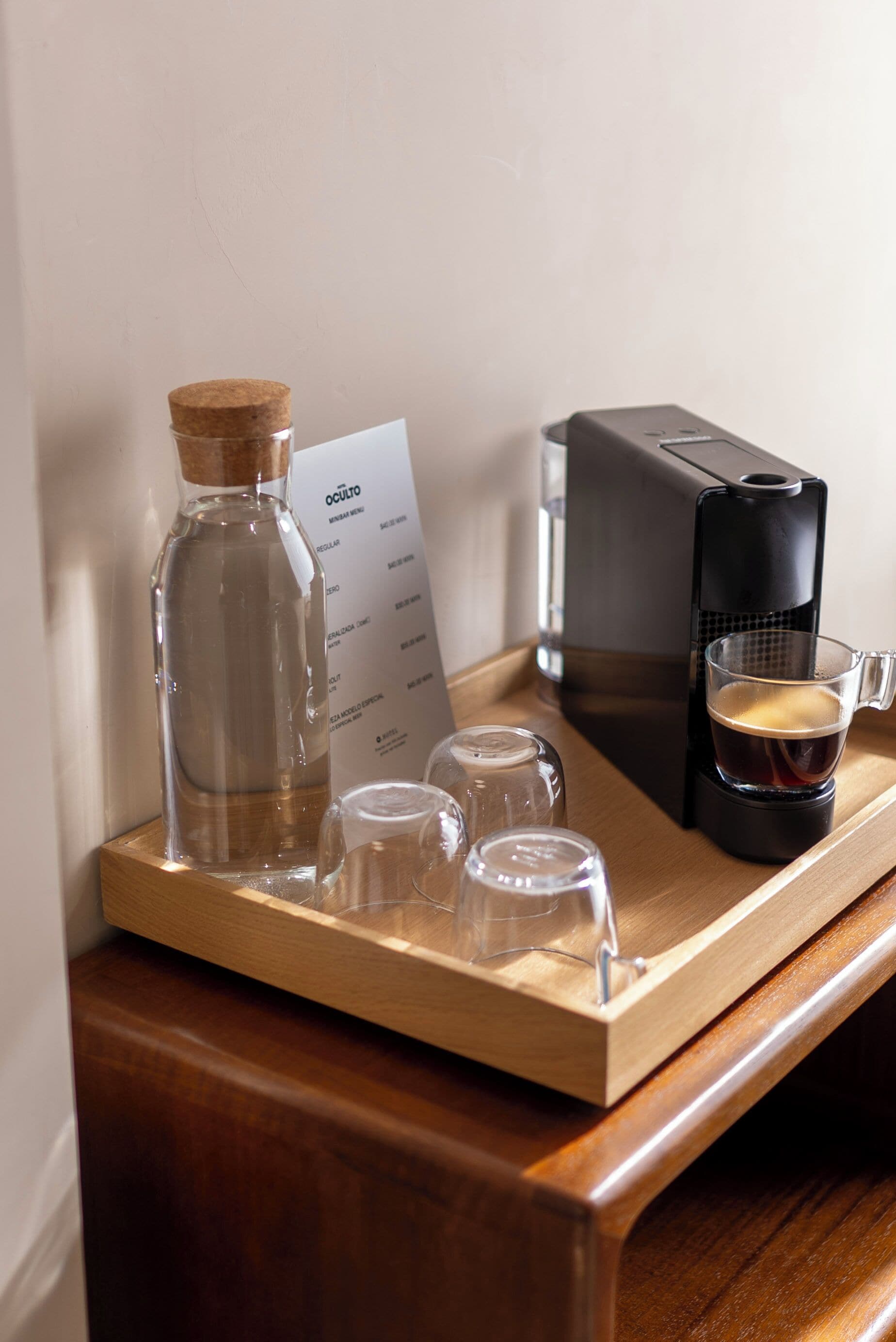 A wooden tray with a coffee maker, a glass of coffee, a bottle of water, and a menu.