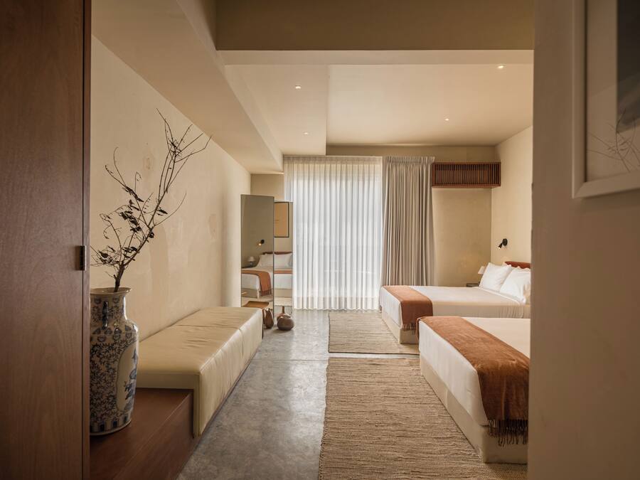 Una habitación de hotel moderna con sofá, dos camas, un ventanal con cortinas, un florero con ramas y un espejo.