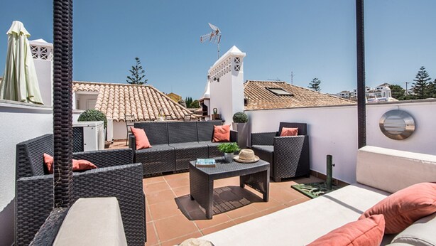 Une terrasse extérieure avec des meubles en osier, une table et un mur blanc en arrière-plan.