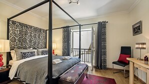 Un lit à baldaquin avec un banc, un bureau et une chaise dans une chambre d’hôtel dotée d’un papier peint à motifs et d’un balcon.