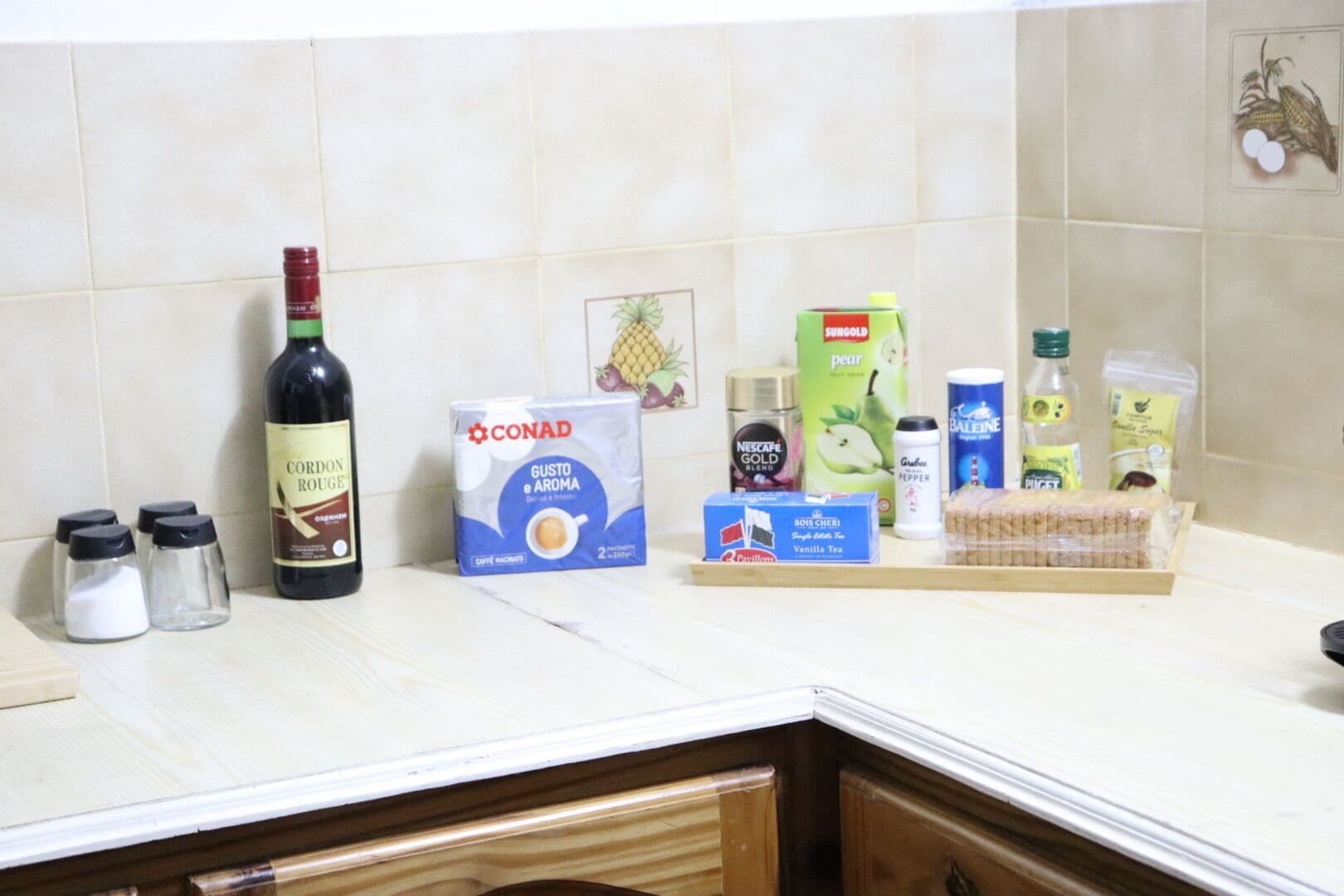 A kitchen counter with various items including a bottle of Cordon Rouge, a box of Conad Gusto e Aroma, a Nescafe Gold Blend coffee, a box of Bois Chéri Vanilla Tea, and a loaf of bread.