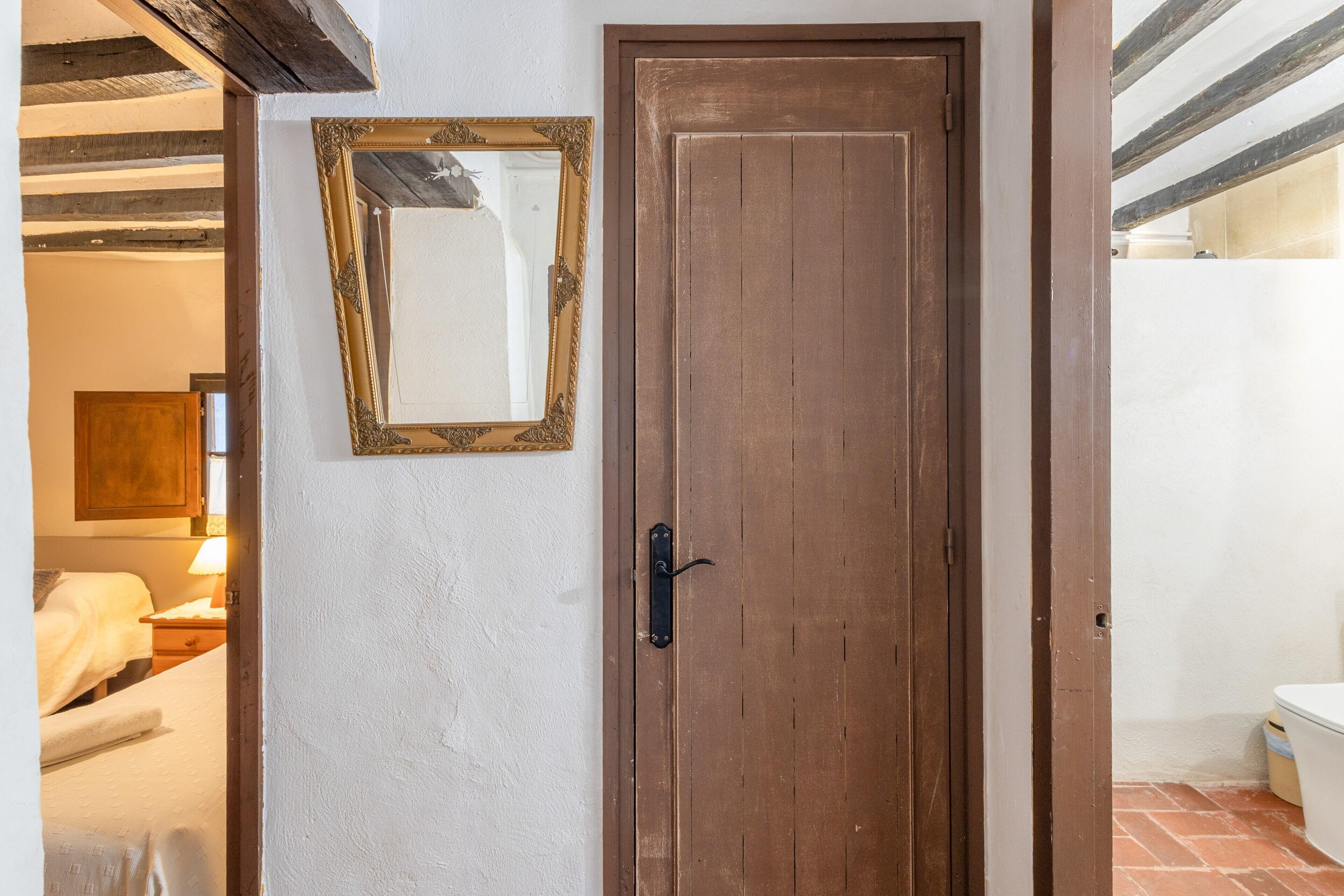 A wooden door with a black handle, a mirror with an ornate gold frame, and a bed with a white cover.