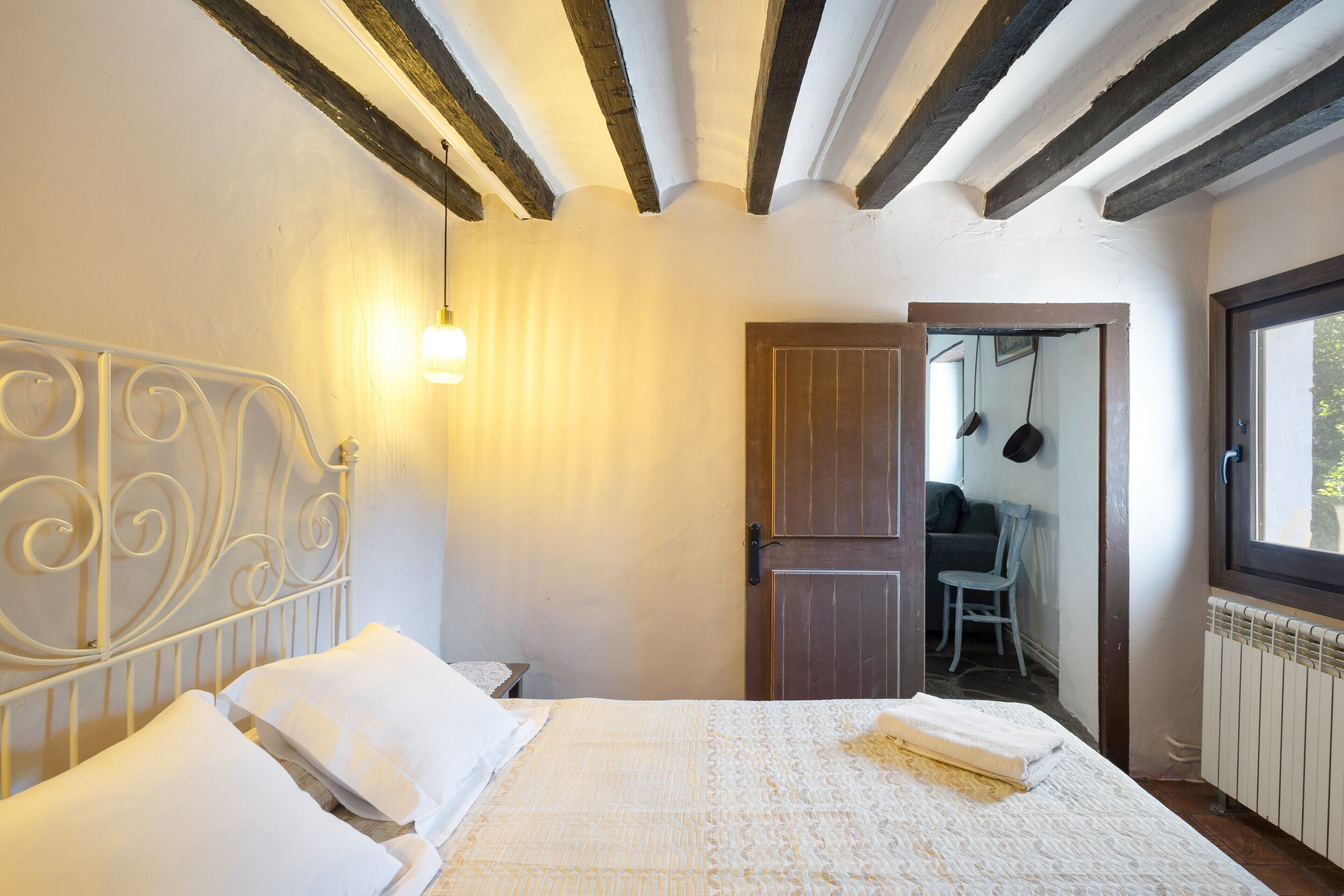 Un dormitorio con cama, almohadas blancas, cabecero, luz empotrada en la pared, puerta de madera y ventana.
