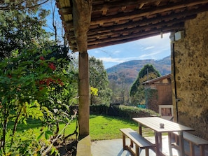 A rustic wooden pergola with a table and benches, a stone wall, and a view of a green garden and hills. - Granota de Mas Violella Country House: Natural Park, Views, Pool & Wi-Fi (Sant Joan les Fonts)