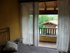 A room with a bed, a small table, lace curtains, and a balcony with a wooden railing. - Granota de Mas Violella Country House: Natural Park, Views, Pool & Wi-Fi (Sant Joan les Fonts)