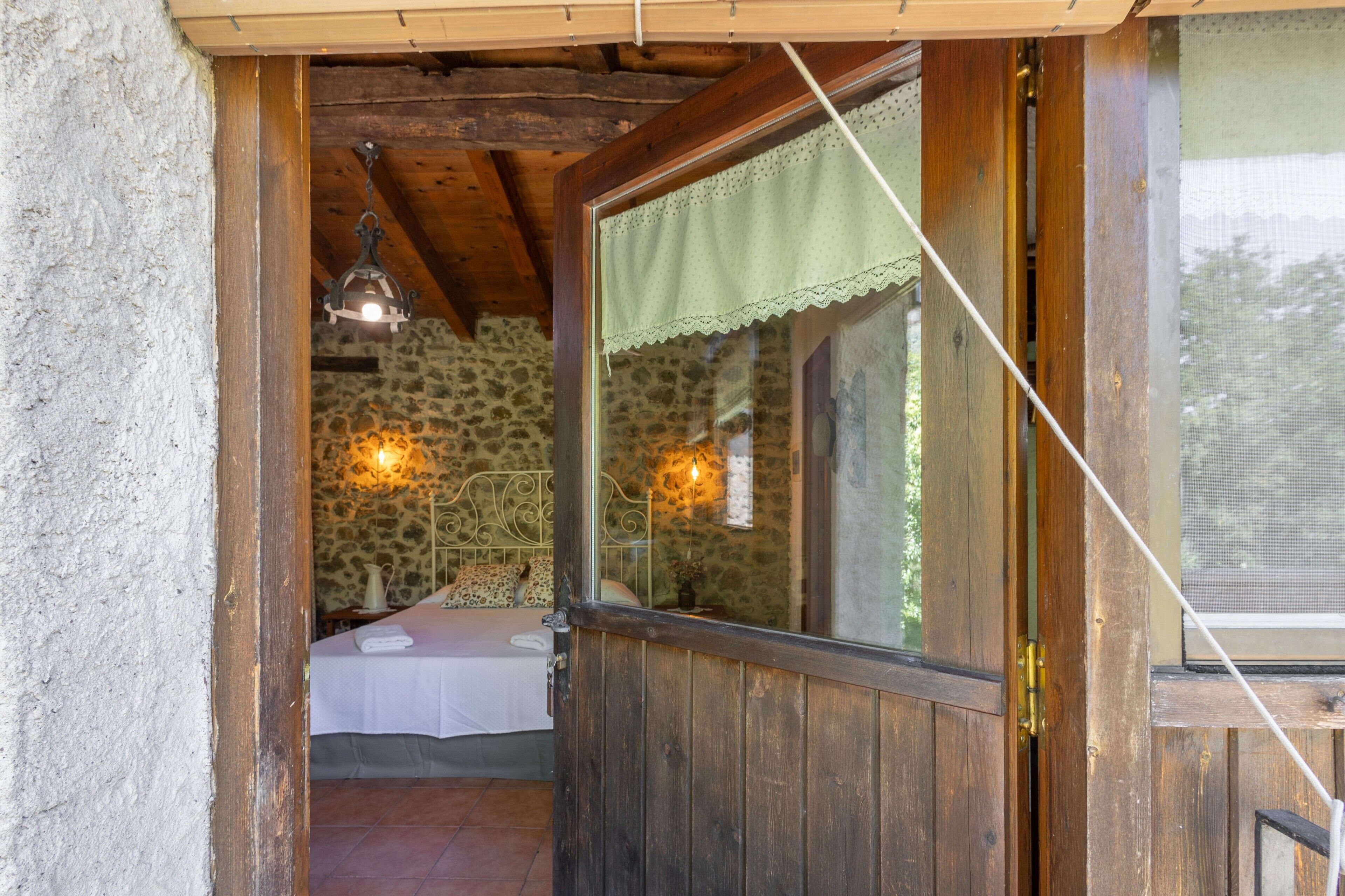 Eine rustikale Holztür führt in einen Schlafzimmerraum mit einer Steinwand, einem Bett mit weißer Bettdecke und einer hängenden Leuchte.