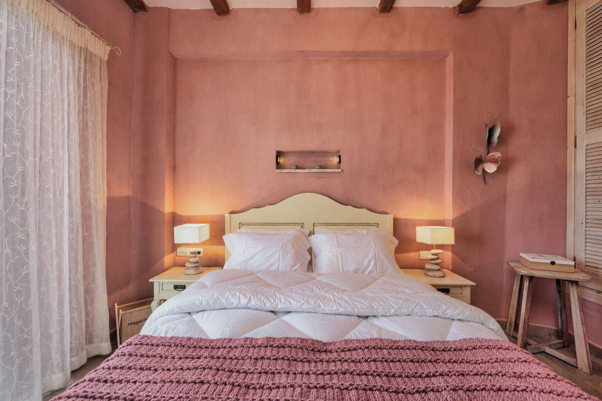 A bedroom with a bed, two bedside tables with lamps, a wooden stool, and a wall-mounted shelf.