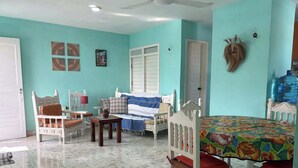 A room with teal walls, a white ceiling fan, a dining table with a floral tablecloth, a sofa with a blue blanket, and a door with a decorative item hanging on it. - Delightful Beach Time in Chelem! (Chelem)