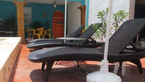 Terraço com cadeiras de descanso e uma mesa, guarda-sol e um edifício com paredes azuis e laranja.