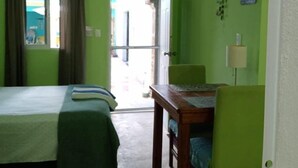 Quarto com duas camas, uma mesa e uma cadeira. Tem uma janela com cortinas, uma porta que dá para o exterior e um ventilador de teto.