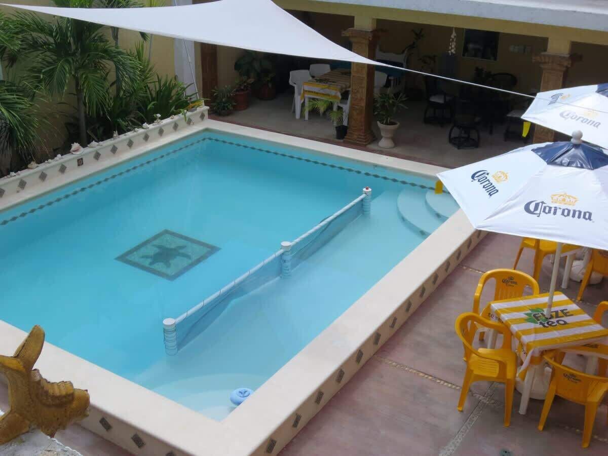 A swimming pool with a lifeguard chair and a table with chairs, surrounded by umbrellas and potted plants.