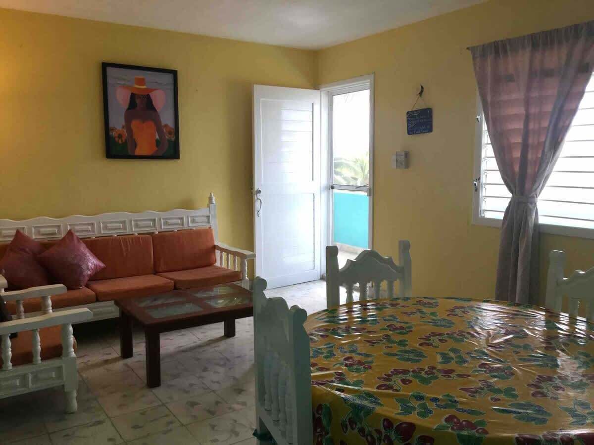 A living room with a sofa, a coffee table, and a dining table with a floral tablecloth. There is a painting on the wall and a door leading outside.
