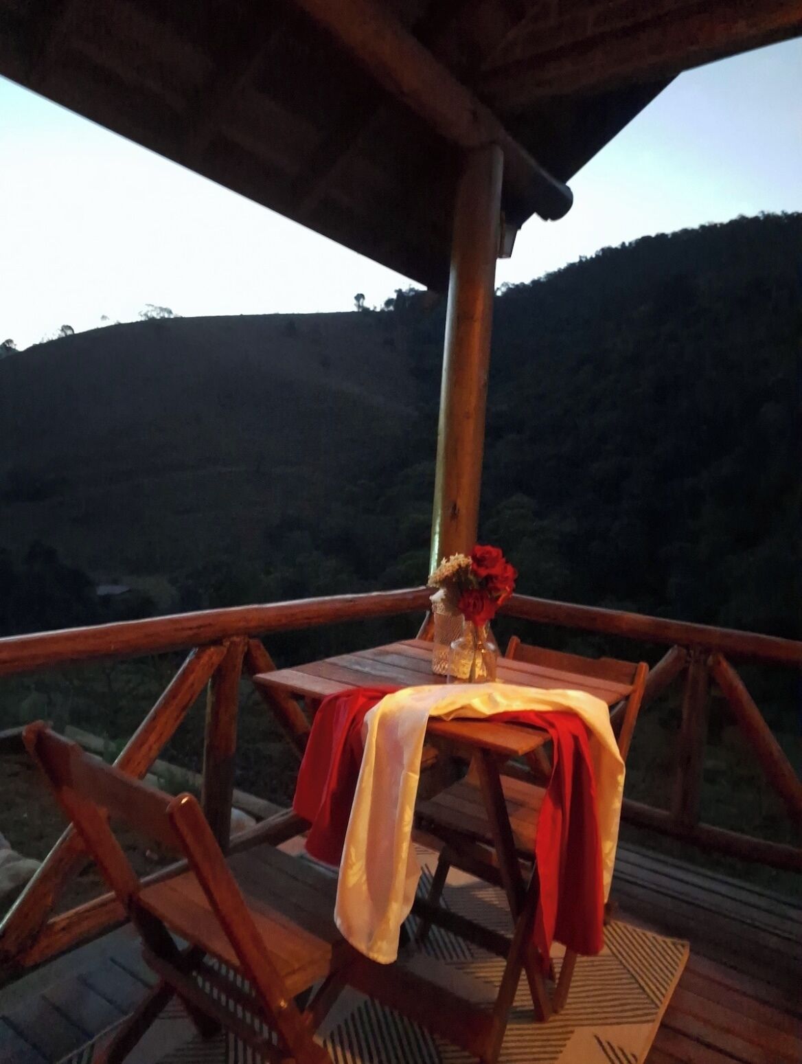 An outdoor wooden deck with a table set for two, a vase with flowers, and a view of a hilly landscape.