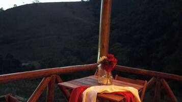 An outdoor wooden deck with a table set for two, a vase with flowers, and a view of a hilly landscape.