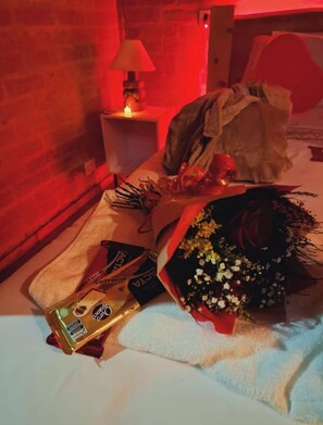 Une pièce faiblement éclairée, avec un lit, une table de chevet sur laquelle se trouve une lampe, et un bouquet de fleurs.