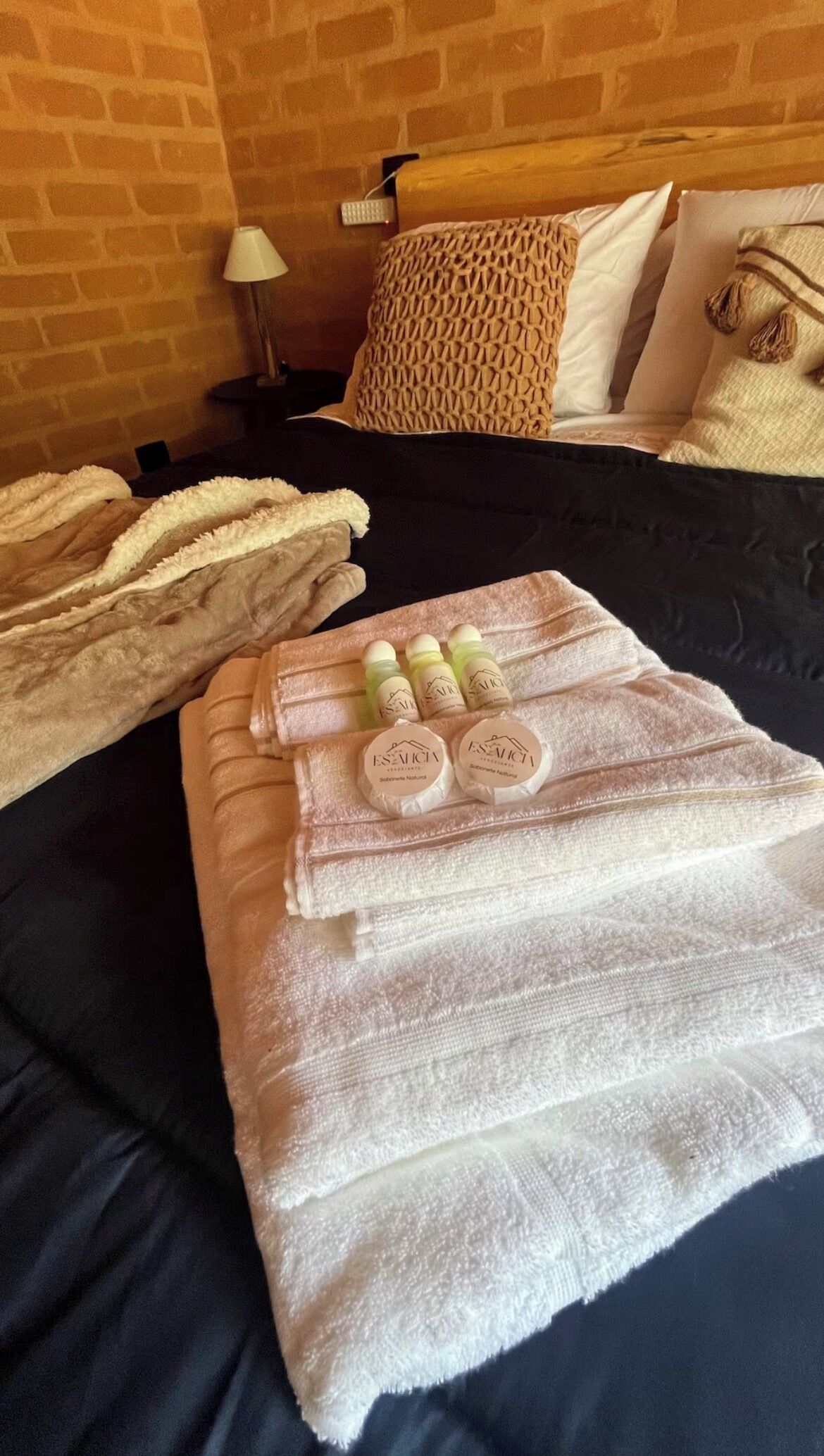 A neatly made bed with a black bedspread, a woven throw blanket, and a set of toiletries including two bottles of shampoo and conditioner, and a bottle of lotion.