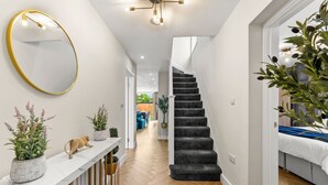Un pasillo moderno con una consola de mármol, plantas en macetas, un espejo con marco dorado, una escalera con alfombra oscura y vistas a un salón.