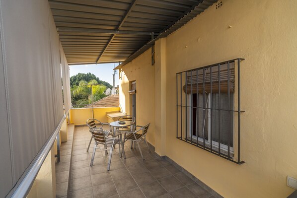 A covered balcony with a table and chairs, a window with bars, and a view of greenery.