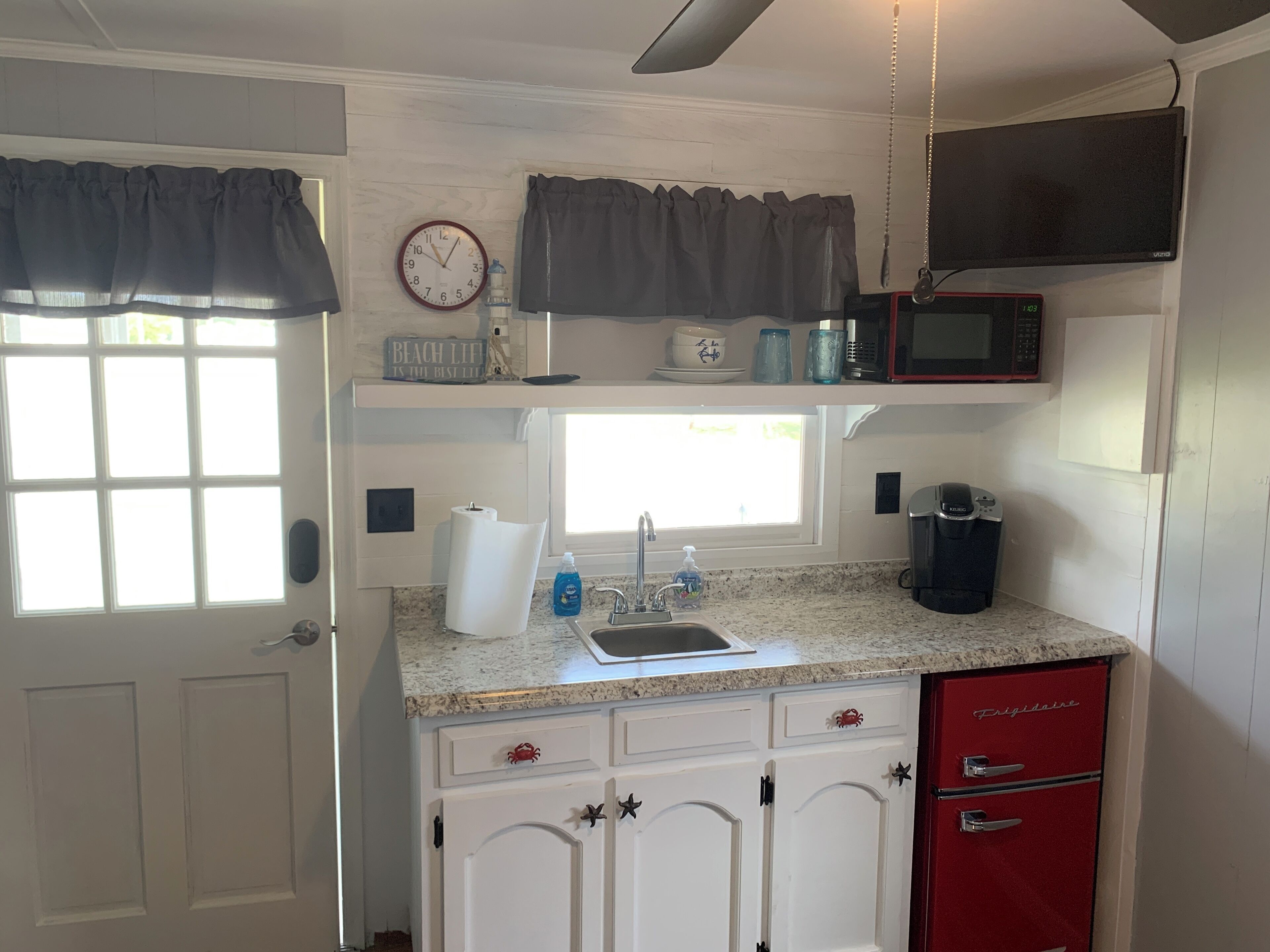 Una cocina compacta con armarios blancos, un refrigerador rojo, un microondas y una ventana con cortinas.