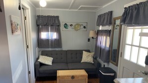 A living room with a grey sofa, a wicker chest, a trash bin, and a kitchen counter. - Cozy cottage (Chincoteague)