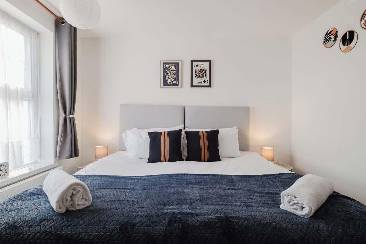 Habitación de hotel con cama, dos lámparas de noche, ventana con cortinas y dos cuadros en la pared.