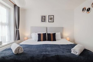 A hotel room with a bed, two bedside lamps, a window with curtains, and two framed pictures on the wall. - Comfortable 3 Bedroom Home Sleeps 5 (Wellingborough)