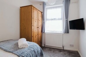 Une chambre à coucher comprenant un lit, une armoire en bois, une fenêtre avec des rideaux, un radiateur et un téléviseur fixé au mur.