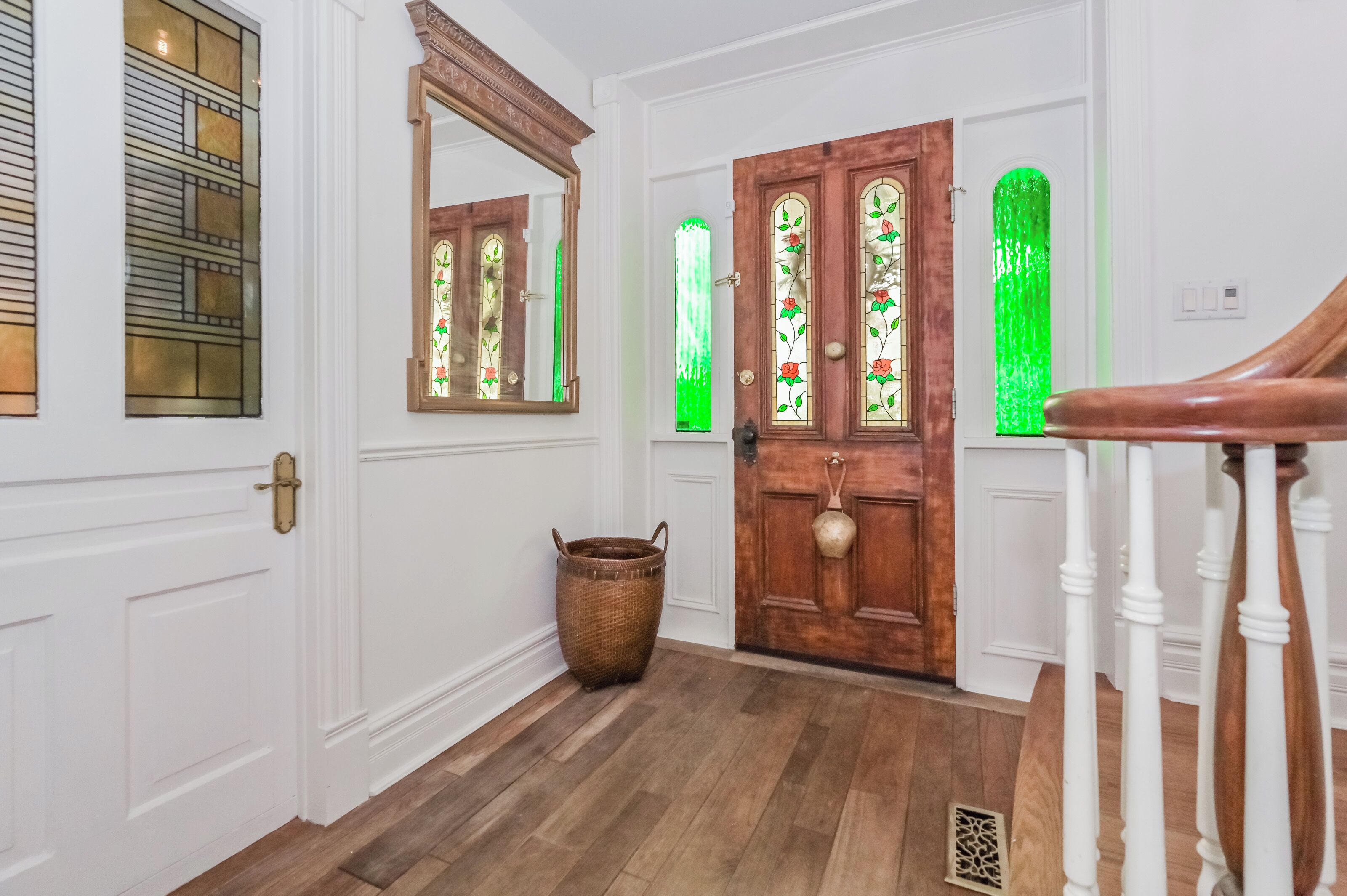 Un pasillo con paredes blancas, suelo de madera, un espejo grande, una puerta de madera con vidrieras y una cesta de mimbre.