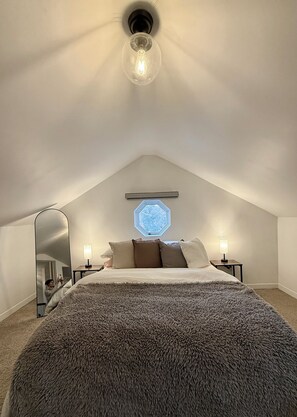 Un dormitorio con techo inclinado, una lámpara redonda, una cama con manta gris suave, dos lámparas de noche y un tragaluz hexagonal.