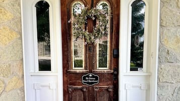 Una puerta de madera con un adorno de laurel, un felpudo y dos macetas con flores.