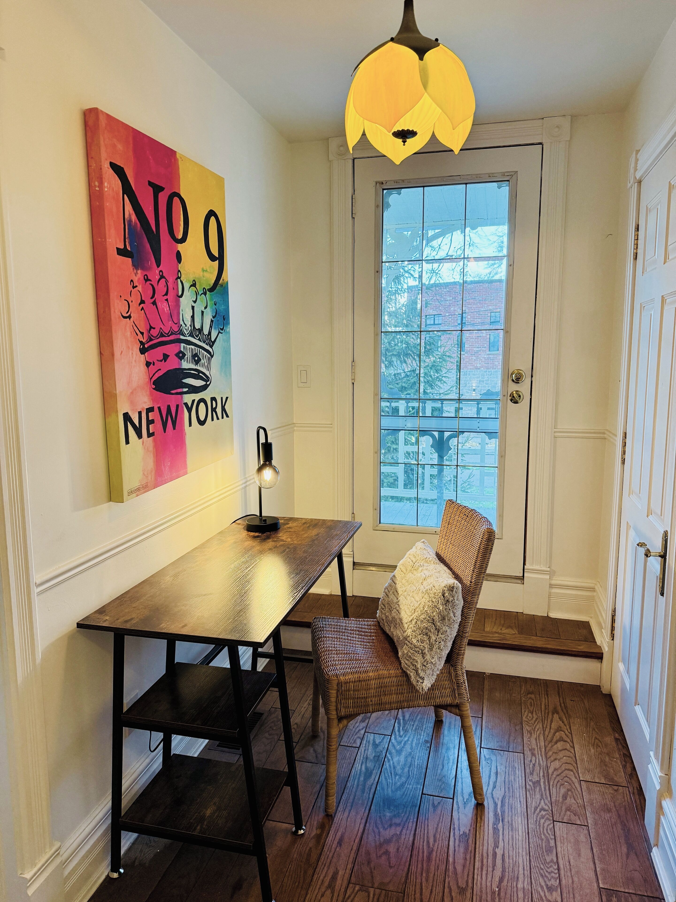 Uma sala de jantar com uma mesa de madeira, uma cadeira, uma luminária pendente e uma janela com vista para edifícios.