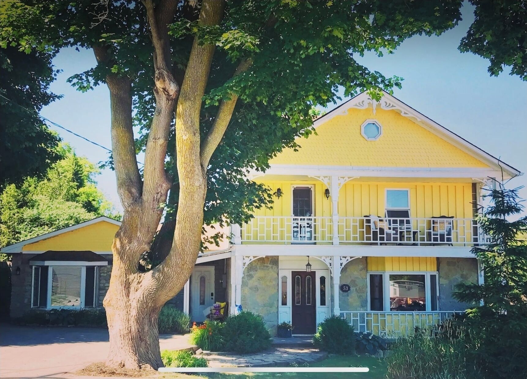 Uma casa amarela de dois andares com varanda, uma casa de pedra ao lado, e uma árvore grande em primeiro plano.