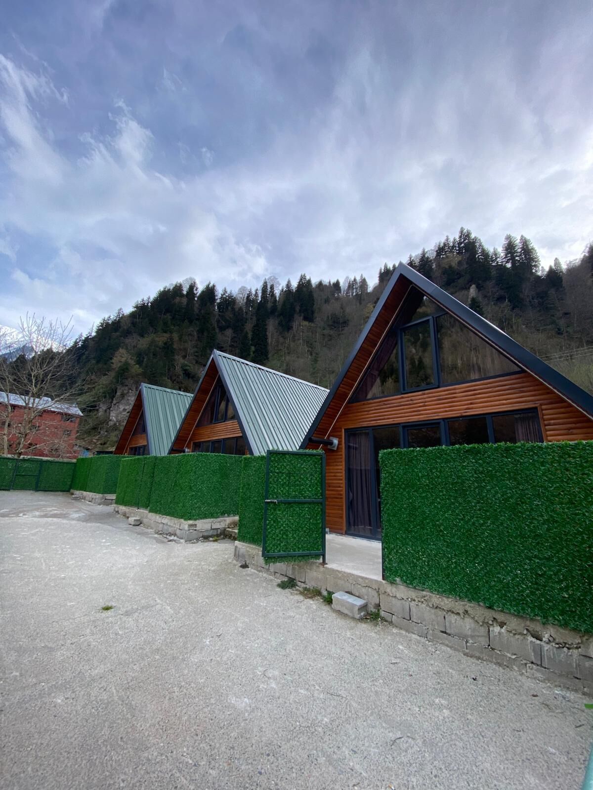 A row of modern cabins with sloped roofs, surrounded by green hedges and set against a mountainous backdrop.
