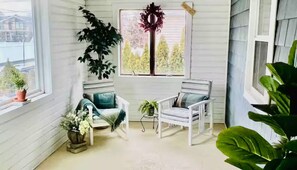 Ein gemütliches Zimmer mit weißen Wänden und einem großen Fenster. Zwei weiße Stühle, ein kleiner Tisch mit einer Pflanze und ein Topf auf der Fensterbank.