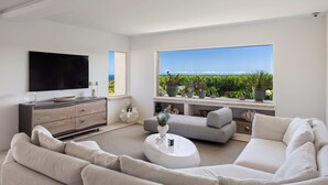 Un soggiorno moderno con una grande TV a schermo piatto, un divano grigio, un tavolino da caffè bianco e vista su un vigneto attraverso un'ampia finestra.