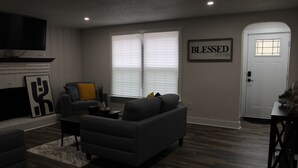 Ein Wohnzimmer mit grauem Sofa, einem Couchtisch und einem Fernseher. Eine Tür mit Fenster und ein Schild mit der Aufschrift 'Blessed'.