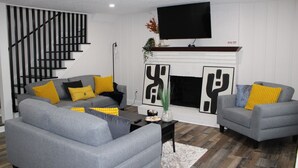 Ein modernes Wohnzimmer mit grauen Sofas, gelben Kissen, einem schwarzen Couchtisch, einem Kamin und einem darüber montierten Fernseher.