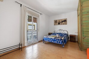 Un dormitorio con cama, mesita de noche, ventana con cortinas, puerta y un cuadro colgado en la pared.