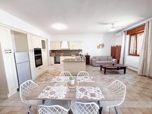 A modern kitchen with white cabinets, a dining area with patterned placemats, and a living room with a sofa and a coffee table. - Villetta Jaclin - Salento Reservation (Taviano)