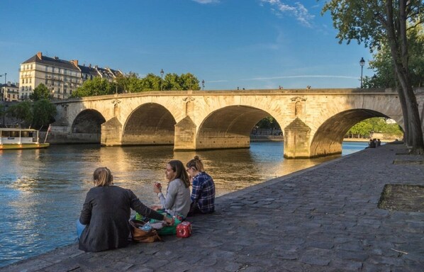 A stone bridge over a river with people sitting on the riverbank. - LUXURIOUS PARISIAN DUPLEX WITH SEINE VIEWS IN HISTORIC ÎLE SAINT-LOUIS - 3 BEDS (Paris)