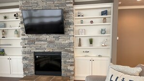 Ein Wohnzimmer mit Kamin aus Stein, einem darüber montierten Flachbildfernseher und einem grauen Sofa mit einem Kissen, auf dem 'Ich liebe See' steht.