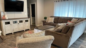 Un salon avec un canapé marron, un fauteuil à motifs et une table basse. Un téléviseur à écran plat est fixé au mur, et il y a un meuble avec des portes en verre.