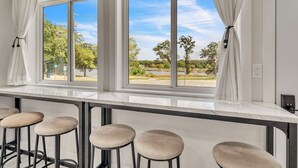 A bar area with a long counter and multiple stools, large windows with curtains, and a view of a lake and trees. - Cozy 3 bedroom Cabin- Close to splash pad (Spirit Lake)