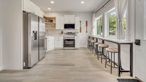 A modern kitchen with white cabinetry, a stainless steel refrigerator, a built-in oven, and a microwave. There are bar stools along the window and a small kitchen island with a sink. - Cozy 3 bedroom Cabin- Close to splash pad (Spirit Lake)