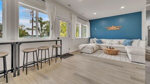 A modern living room with a blue accent wall, a white sofa, a wooden coffee table, and a bar area with stools. - Cozy 3 bedroom Cabin- Close to splash pad (Spirit Lake)