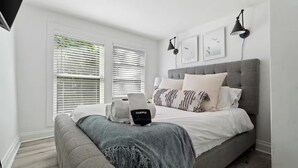 Un dormitorio con una cama tapizada en gris, ropa de cama blanca y una manta gris. Hay dos lámparas negras fijadas a la pared, un cabecero gris y una ventana con persianas.