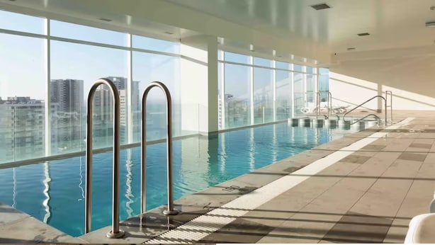 Una piscina interior con paredes de vidrio transparente, escaleras de metal y suelo de baldosas.