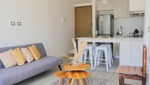 Una sala de estar moderna con un sofá gris, una mesita de madera con un jarrón, y una zona de cocina con refrigerador, taburetes de bar y una silla de bambú.